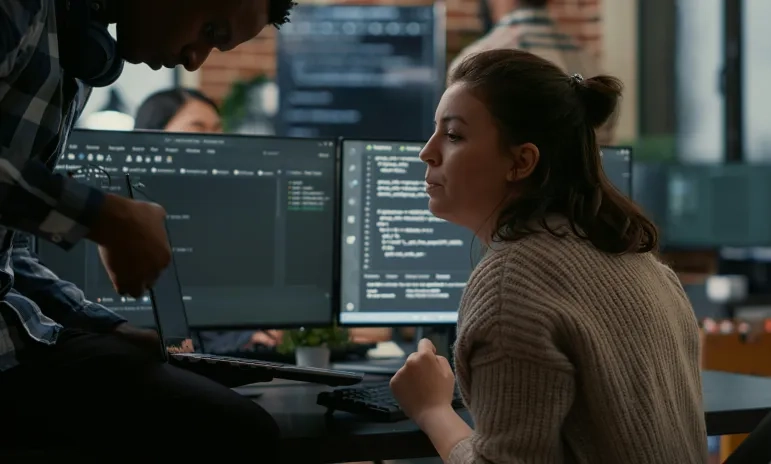 Two coworkers collaborate at a desk in a modern office, reviewing code on dual computer monitors while one person holds a laptop and the other looks on attentively.