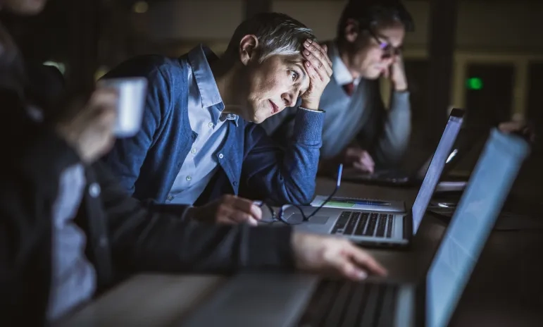 Tired office workers working late at laptops, one person holding their head in frustration.