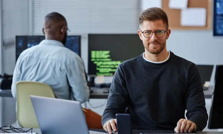A developer smiling at the camera in an open-plan office, seated at a desk with a laptop, with a colleague working at a monitor displaying green code in the background.