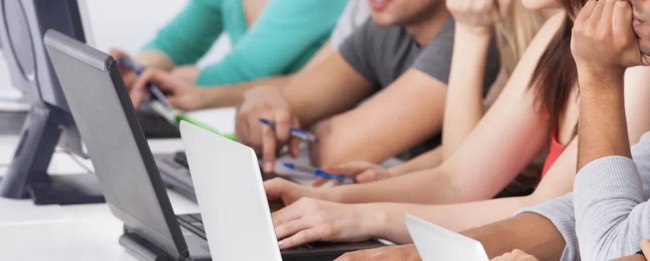 A group of students sitting in a row, working on laptops and taking notes in a classroom setting.