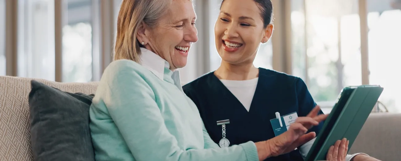 A smiling healthcare professional in a navy uniform assists a happy elderly woman with a tablet while sitting on a couch in a bright, modern setting.
