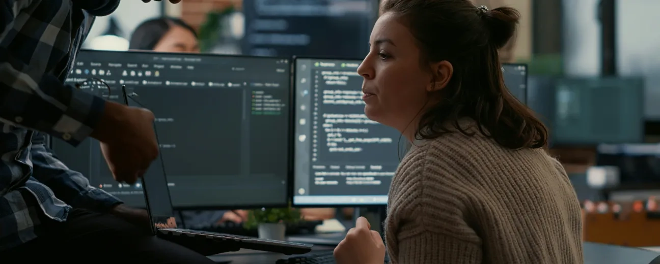 Two coworkers collaborate at a desk in a modern office, reviewing code on dual computer monitors while one person holds a laptop and the other looks on attentively.