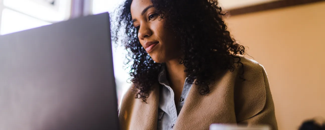 A woman with curly hair sits at a café table, focused on her laptop screen while holding a coffee mug, embodying mindful and sustainable web design practices.