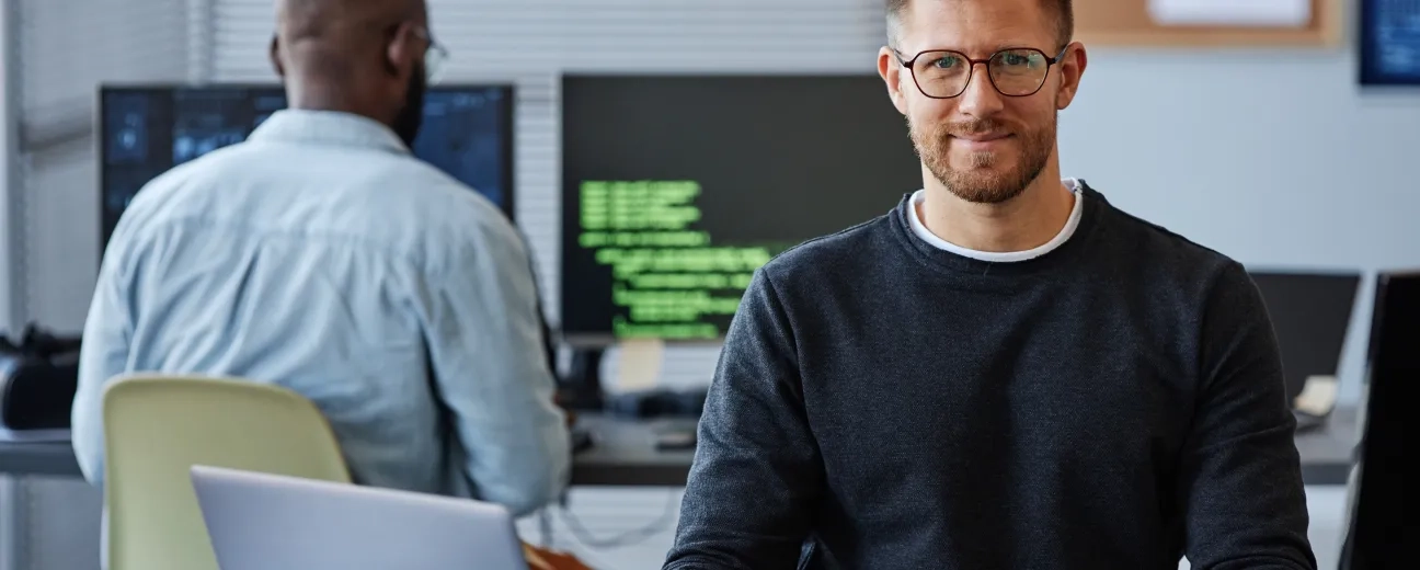 A developer smiling at the camera in an open-plan office, seated at a desk with a laptop, with a colleague working at a monitor displaying green code in the background.