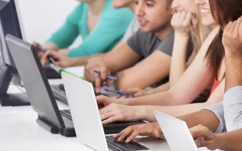 A group of students sitting in a row, working on laptops and taking notes in a classroom setting.