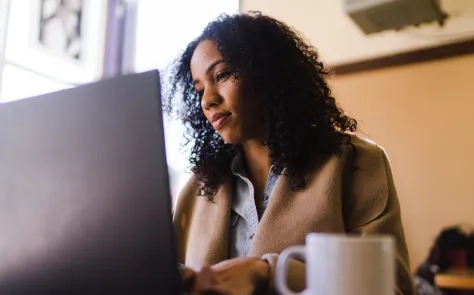 A woman with curly hair sits at a café table, focused on her laptop screen while holding a coffee mug, embodying mindful and sustainable web design practices.