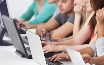 A group of students sitting in a row, working on laptops and taking notes in a classroom setting.